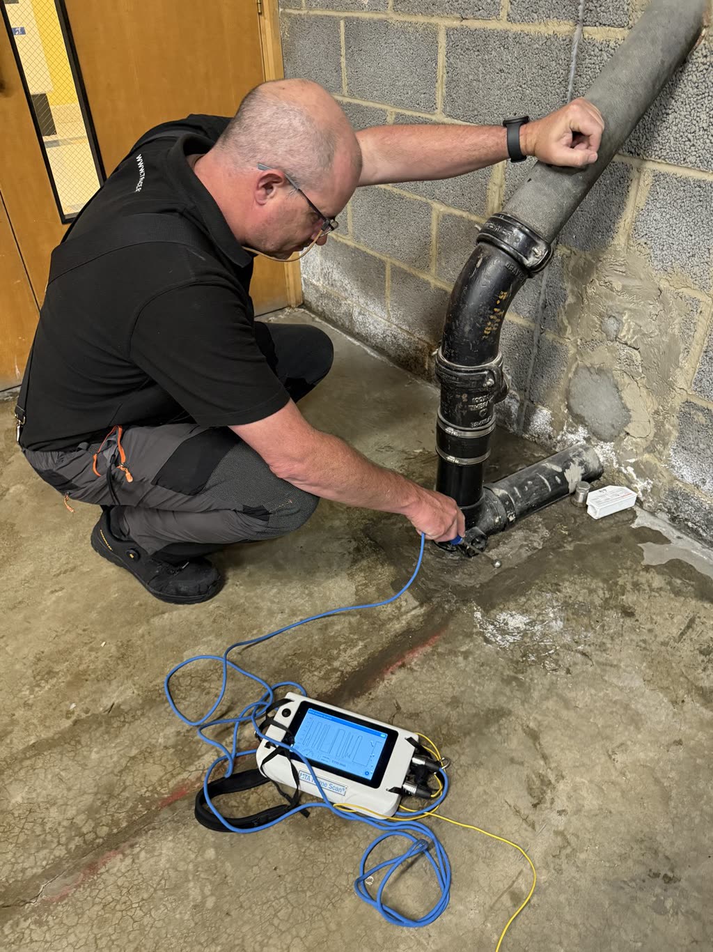 A Trace Surveys surveyor testing beside a pipe connection at floor level with a portable instrument.